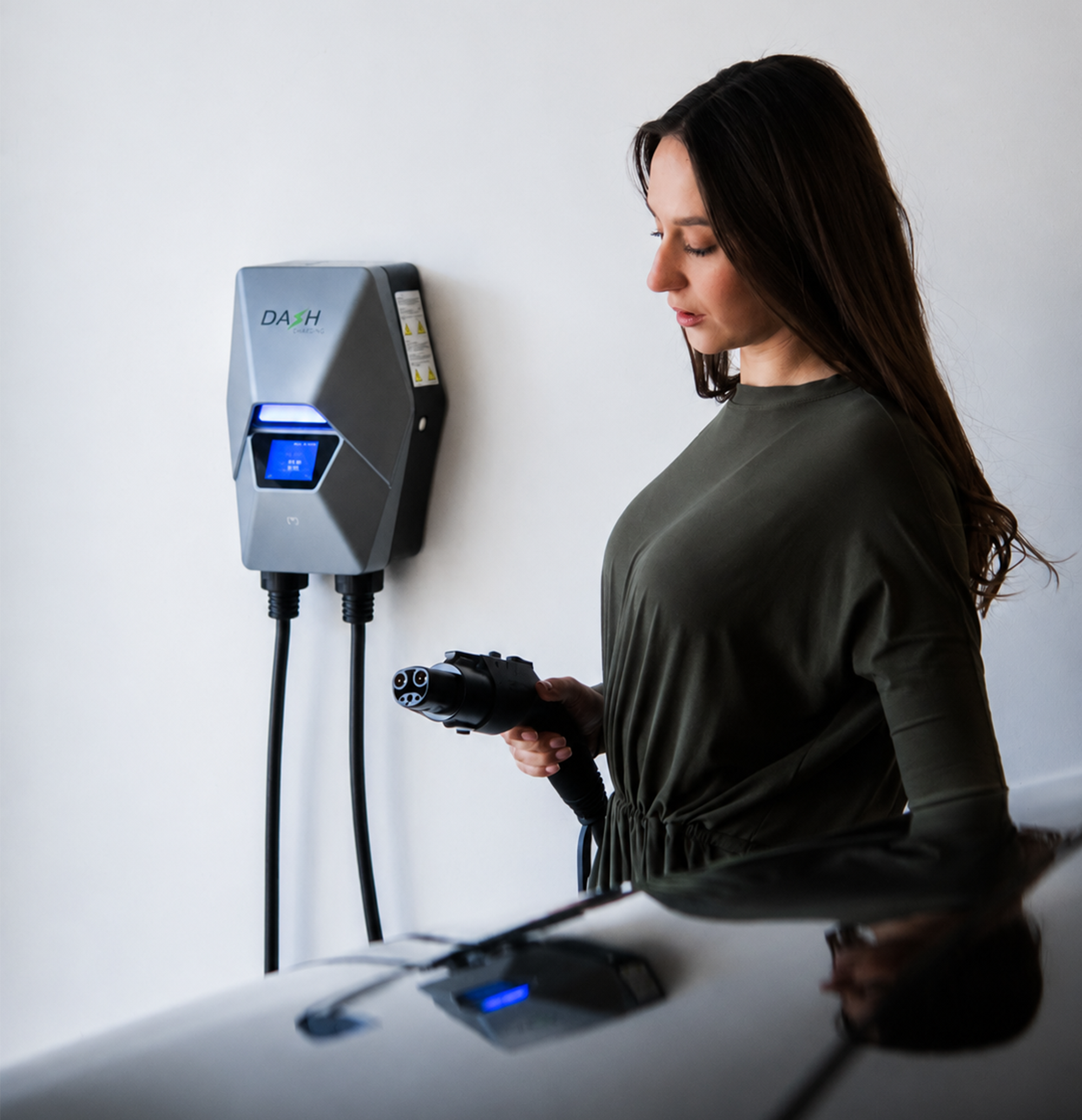 A woman in a dark olive green dress is shown from the side as she plugs an electric vehicle charger into a car. The wall-mounted charger is gray with a blue digital display screen, mounted on a plain white wall.